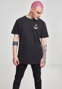 Černé bavlněné tričko s krátkým rukávem, s motivem zlatého kotvy na levé části hrudi. Model má na sobě černé roztrhané džíny.
