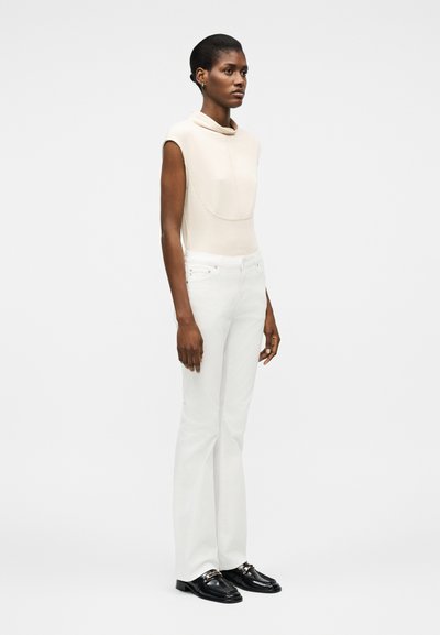 Γυναίκα που στέκεται κοιτώντας μπροστά, φοράει μπεζ τοπ χωρίς μανίκια, λευκό παντελόνι και μαύρα μοκασίνια, μπροστά σε απλό λευκό φόντο.