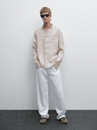 Beige Leinenhemd mit langen Ärmeln und Brusttasche, kombiniert mit weißen lockeren Hosen und grauen Schuhen. Flache Pose vor einer schlichten Wand.