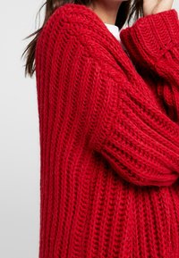 Cárdigan de punto rojo que presenta una textura gruesa y acanalada, con mangas de gran tamaño y un corte relajado. Enfocado en la calidez y la comodidad en su diseño.