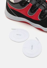 Zapatilla negra y roja con parte superior de malla, suela gris y cordones negros. Dos cordones redondos blancos están colocados al lado de la zapatilla.