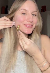 Rouleau facial en or tenu contre la joue, avec des extrémités arrondies et une finition métallique. Le modèle affiche une peau claire et porte des bijoux minimalistes.