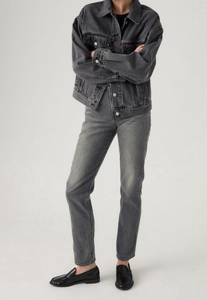 Person iført mørkegrå denimjakke og jeans med sorte loafers, stående med armene krydset mod en ensfarvet baggrund.