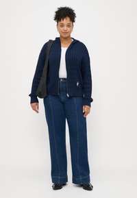 Σκούρο μπλε πλεκτό φούτερ με υφή, σε συνδυασμό με ψηλόμεσα μπλε τζιν. Το μοντέλο κρατάει μια μαύρη τσάντα. Απλός σχεδιασμός και άνετη εφαρμογή.