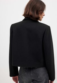 Schwarze, kurz geschnittene Jacke mit strukturiertem Design, langen Ärmeln und glatter Textur. Verfügt über einen klassischen Kragen und minimale Hardware-Akzente.