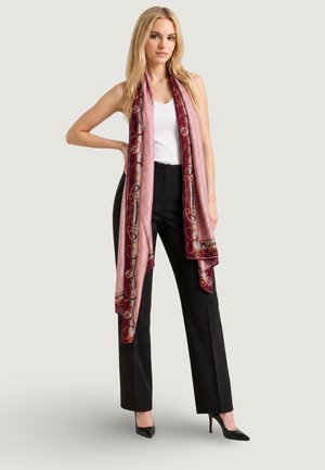 Donna bionda che indossa pantaloni neri, top bianco senza maniche, tacchi neri e una lunga sciarpa rosa e bordeaux con motivi su sfondo grigio chiaro.