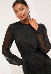 Fekete ruha virágos hímzéssel és átlátszó hosszú ujjakkal, deréknál megkötve. Simogatott textúra és finom részletek mindenhol.