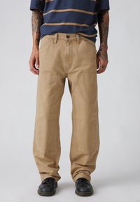 Μπεζ cargo παντελόνι με άνετη εφαρμογή, με μεγάλες μπροστινές τσέπες και ενισχυμένες ραφές. Φορεμένο με μπλε ριγέ πουκάμισο και μαύρα παπούτσια.