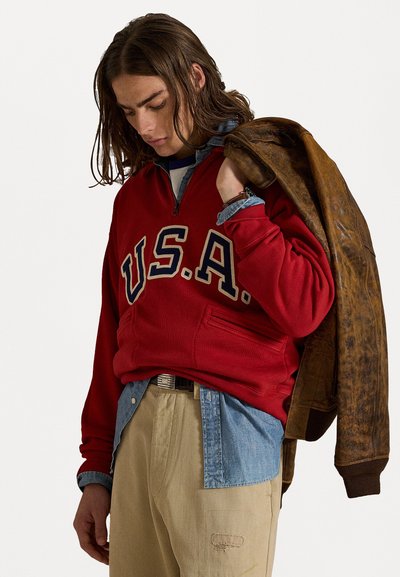 Sweatshirt roșu cu fermoar pe jumătate și inscripția "U.S.A." în albastru, pantaloni khaki și o cămașă albastră din denim în straturi, ținând o jachet�ă maro din piele.