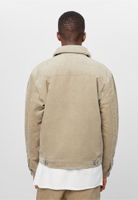 Béžová manšestrová bunda s límcem z jehněčí kožešiny, volného střihu, s knoflíkovými manžetami a knoflíkovými detaily na zádech. Hladký povrch, žebrovaný vzor.