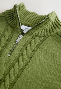 Jersey verde con cremallera, cuello de canalé y detalles de punto con trenzas. Presenta una cremallera plateada y una tela de punto suave y texturizada.