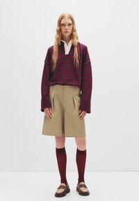 Gestreiftes rotes und marineblaues Langarmoberteil mit einem weißen Kragen, kombiniert mit beigen Shorts und bordeauxroten Kniestrümpfen, ergänzt durch braune Slipper.