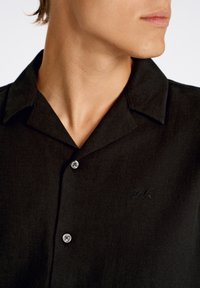 Chemise noire à col, avec un design à boutons, un tissu texturé et un petit logo brodé sur la zone gauche de la poitrine.