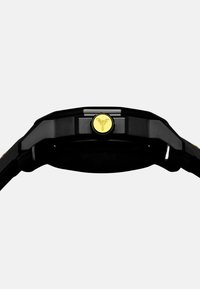 Schwarz Uhr mit einem strukturierten Armband und einer runden, goldenen belüfteten Krone mit einem Logo. Das Gehäuse ist eckig mit polierten Akzenten.