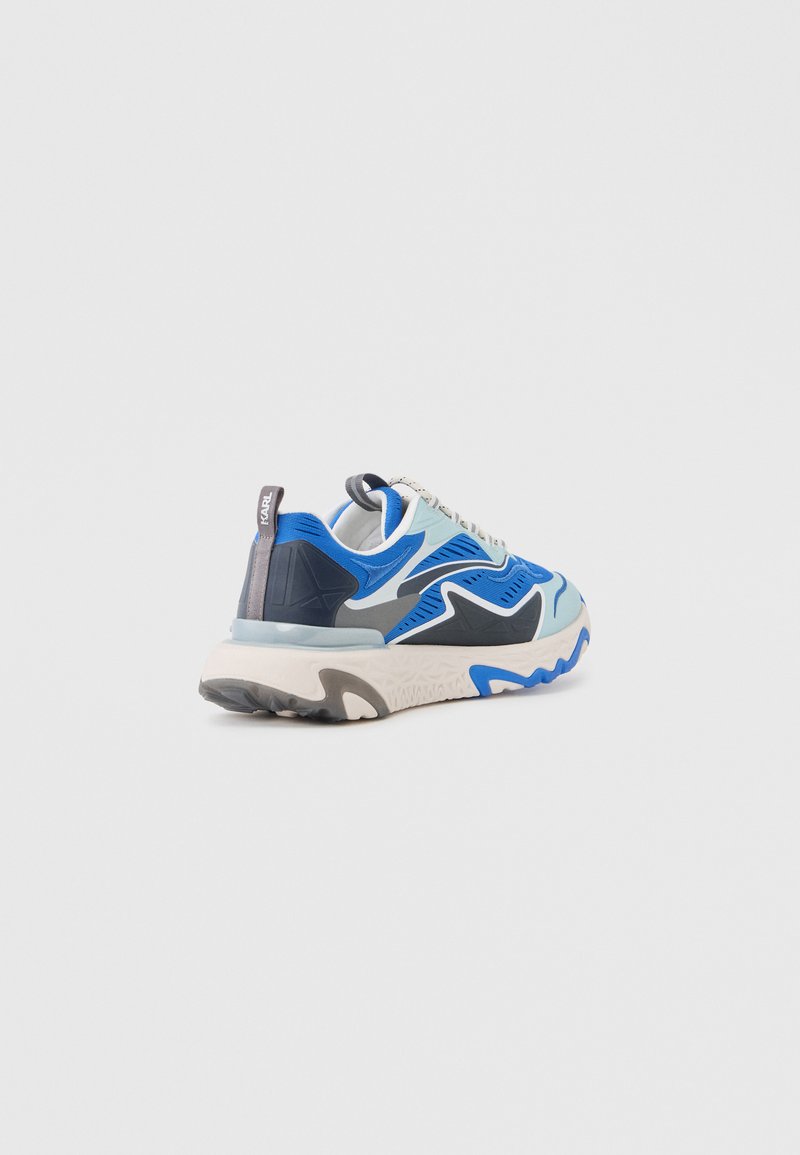 Zapatilla deportiva azul, gris y blanca con patrones ondulados y suela gruesa, vista desde atrás en ángulo.