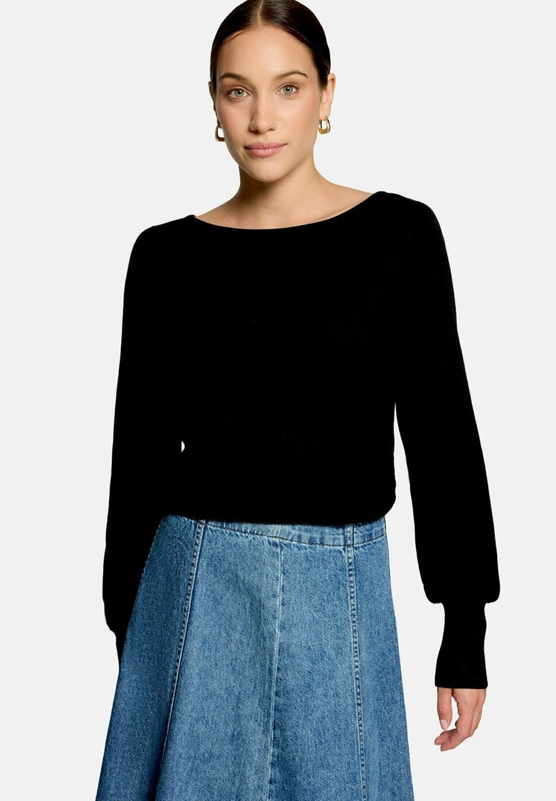 Femme aux cheveux foncés portant des boucles d'oreilles créoles dorées, un haut noir à manches longues et une jupe en jean bleue sur un fond uni.
