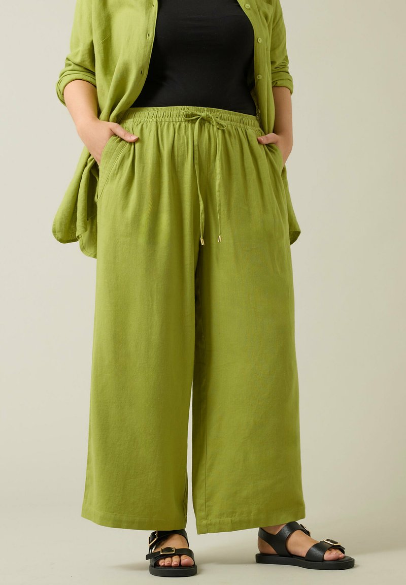 Lichtgroene linnen broeken met wijde pijpen, een elastische tailleband, zijzakken en details met een trekkoord. Gecombineerd met zwarte sandalen met gouden accenten.