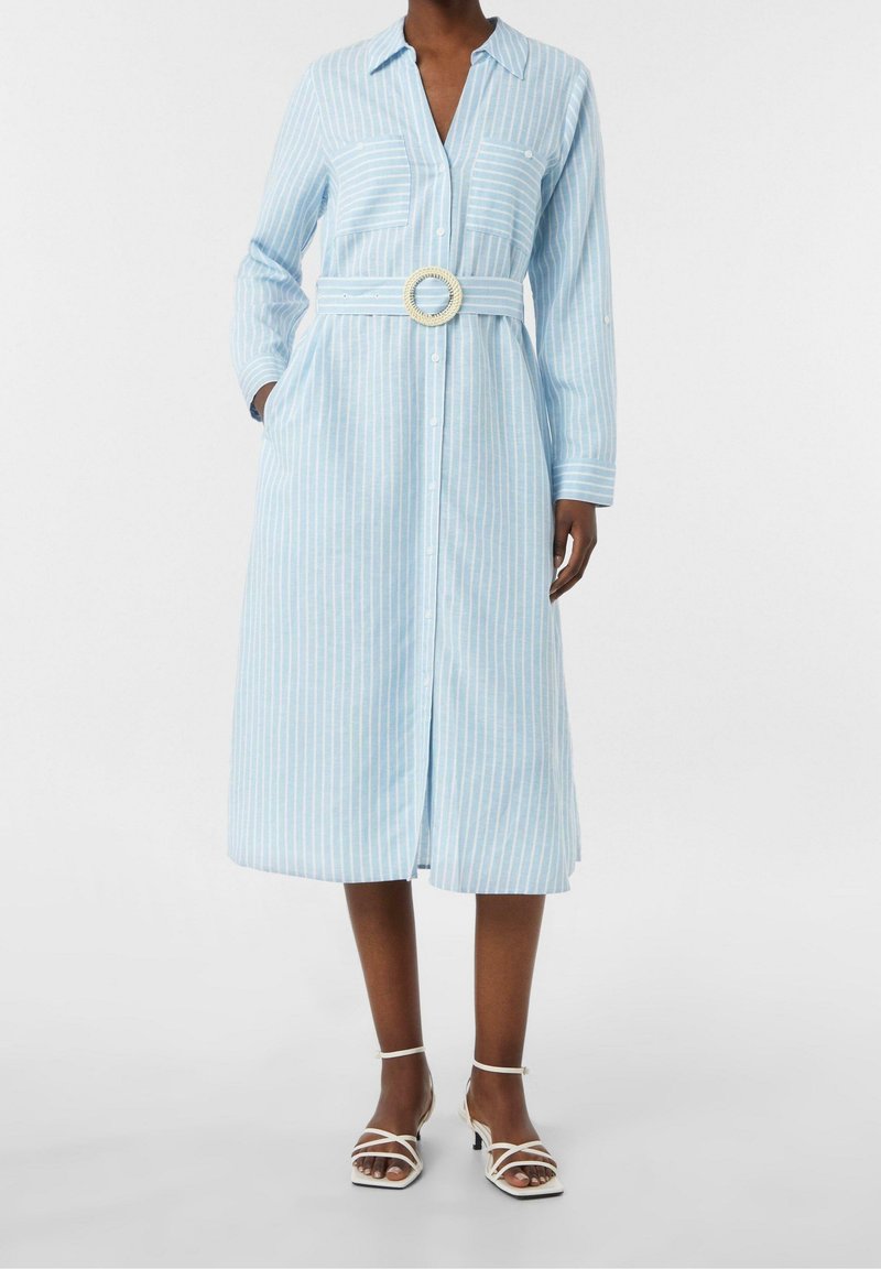 Femme portant une robe midi à rayures bleu clair et blanc à manches longues, avec une boucle de ceinture circulaire et des sandales blanches à talons.