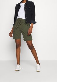 Shorts vert olive avec une taille à cordon, avec des ourlets roulés et des poches latérales, associés à un t-shirt blanc et une veste en denim noire.