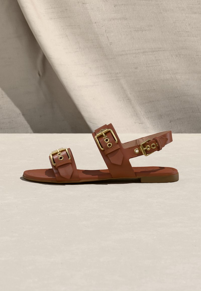 Sandale plate marron avec trois lanières en cuir et boucles dorées sur une surface et un fond clair neutre.