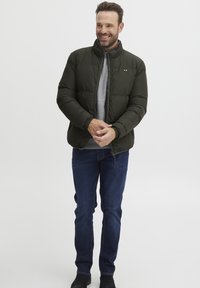 Puffjakke i mørkegrønn, med høy krage, glidelåslukking og elastiske mansjetter. Brukt over en grå skjorte med blå denimjeans.