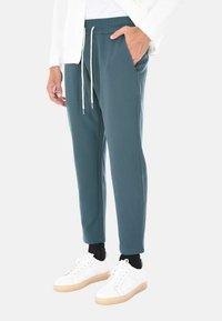 Uomo che indossa pantaloni teal con coulisse, sneakers bianche con suole color marrone chiaro, calzini neri e una giacca bianca con texture.