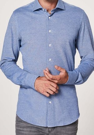 Hombre con una camisa de manga larga azul claro con botones y cuello, ajustándose el puño izquierdo con la mano derecha.