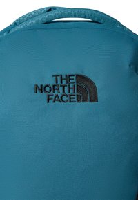 Teal tygsrygga med en strukturerad yta, som har en svart broderad logotyp av "The North Face" nära toppen.