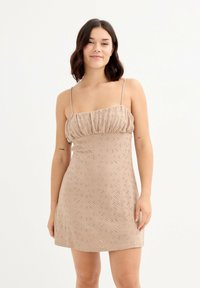 Vestido corto beige con un cuerpo ajustado, escote abombado y tirantes delgados. Presenta un patrón texturizado con recortes en toda la prenda.