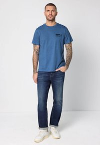 Blå t-shirt med rund hals och svart text "TOUGH TRAILS", kombinerad med mörka jeans och vita sneakers, mot en enkel bakgrund.