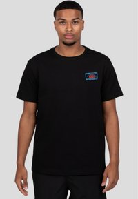 Camiseta de algodón negra con cuello redondo y mangas cortas, que presenta un parche de logo colorido en el pecho. Diseño de corte clásico.