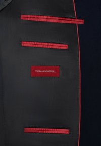 Innenansicht einer marineblauen Stoffjacke mit roten Ribbon-Akzenten und Taschen. Verfügt über ein rotes Etikett mit dem Namen "THOMAS GOODWIN", aufgenäht. Geschmeidige Textur.