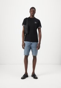 T-shirt preta com detalhes em riscas brancas, calções de ganga com bainhas desgrenhadas e sapatos pretos sem atacadores. Uma pequena mala de ombro preta é usada.