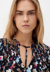 Marinebluse mit floralen Motiven und smockierten Details, ausgestattet mit einem Schlüssel-Ausschnitt und bunten Spritzern in Pink, Blau und Braun auf einem dunklen Hintergrund.