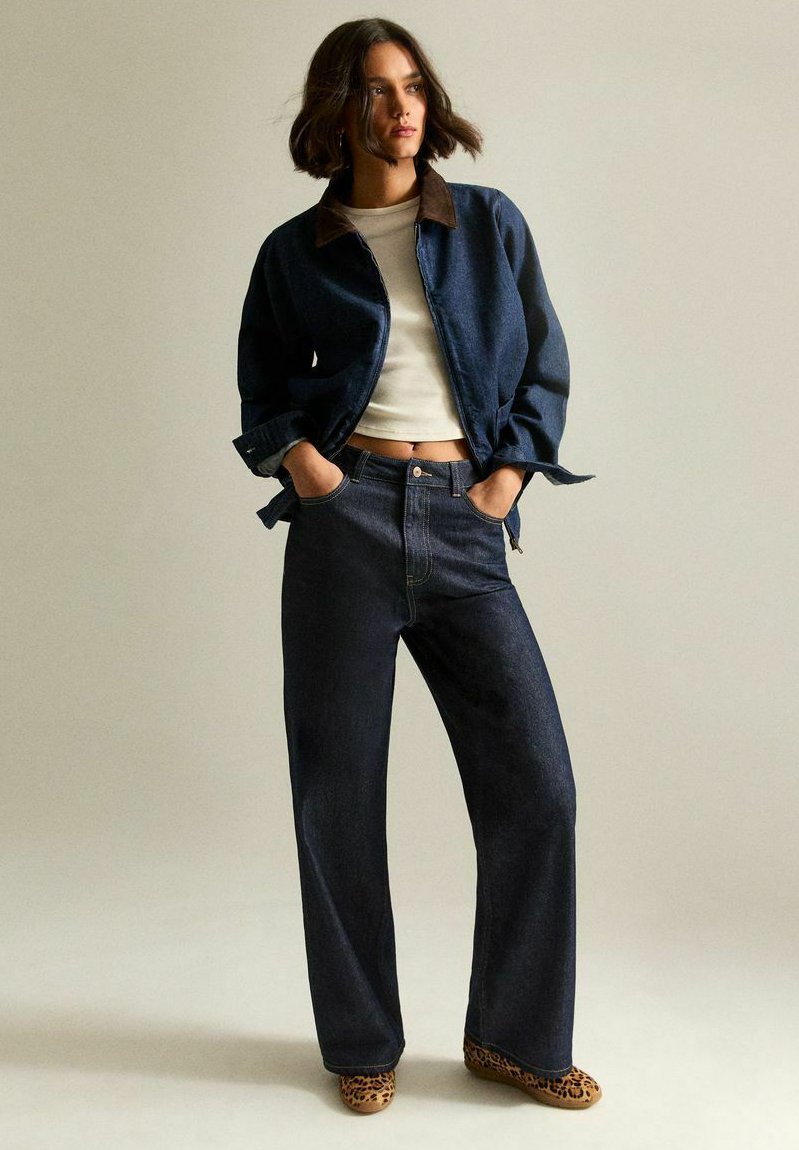 Donna in piedi con le mani in tasca, che indossa una giacca di jeans blu scuro e jeans, una maglietta corta bianca e scarpe con stampa leopardata.