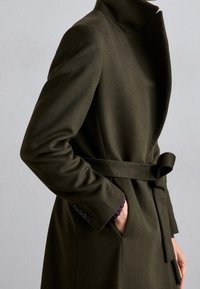 Cappotto di lana verde scuro con colletto alto, vita con cintura e tasche laterali. Presenta una linea dritta e dettagli con bottoni sulle maniche.