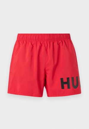 Røde badeshorts med elastisk midje og svart fet delvis «HUG»-tekst på høyre ben, vist mot en lys bakgrunn.