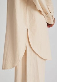 Camicia beige, leggera, con orlo arcuato, polsini con bottoni e maniche lunghe. Tessuto liscio e leggermente stropicciato.