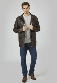 Braune Lederjacke mit Reißverschluss und Knopf-Taschen, kombiniert mit einem hellgrauen Henley-Shirt, blauen Jeans und braunen Schuhen.