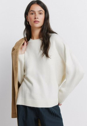 Pitkähiuksinen nainen, jolla on valkoinen neulepaita ja liituraitahousut, pitelee vaaleanruskeaa takkia yhden olkapäänsä päällä yksiväristä taustaa vasten.