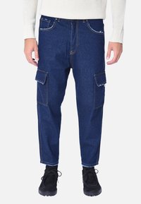 Pantaloni cargo in denim blu scuro con due tasche laterali, cuciture a contrasto e vestibilità affusolata. Indossati con scarpe nere stringate.