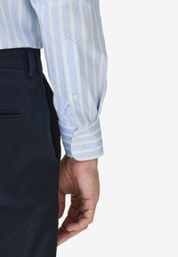 Sininen ja valkoinen raidallinen paidan hiha nappidetaililla, yhdistettynä tummiin kangashousuihin. Ominaisuuksiin kuuluu muototettu malli ja sileä pinta.