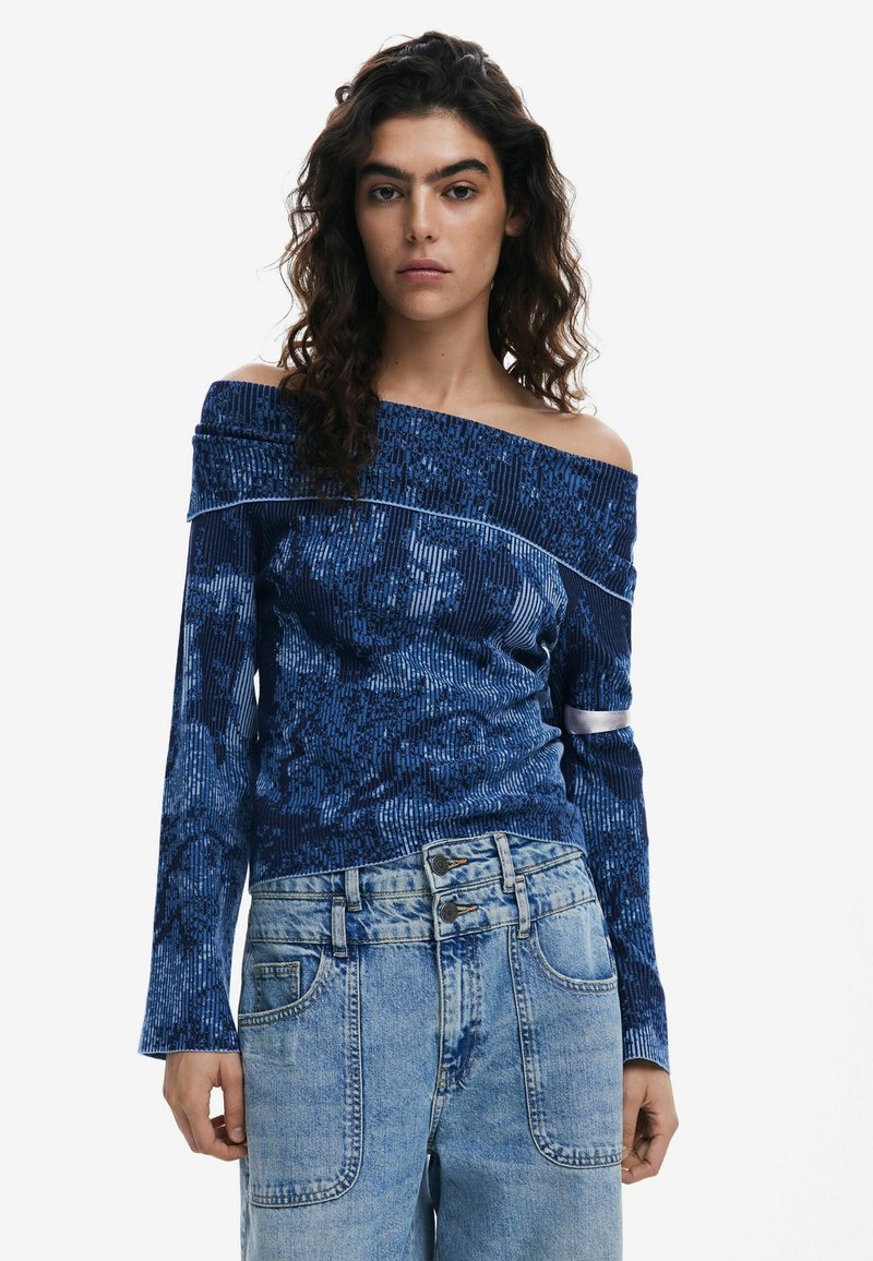 Blaues, schulterfreies Oberteil mit langen Ärmeln, strukturiertem Stoff und kontrastierenden Abschlüssen. Kombiniert mit hellblauen, hoch taillierten Jeans mit doppelten Knöpfen.