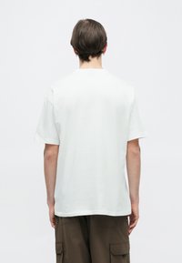 Fehér pamut póló kerek nyakkivágással és rövid ujjakkal. Simább textúrájú, kényelmes szabású, nincs rajta látható minta vagy grafika.