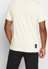 Krämbefärgad kortärmad t-shirt i mjukt tyg. Har en svart etikett längst ner till vänster och en enkel, slät textur.