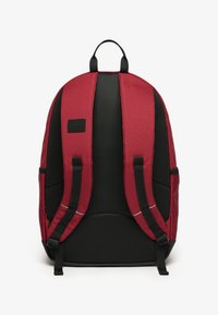 Mochila de tela roja con parte trasera de malla negra, correas acolchadas y un parche de logo. Presenta un diseño ergonómico para mayor comodidad y durabilidad.