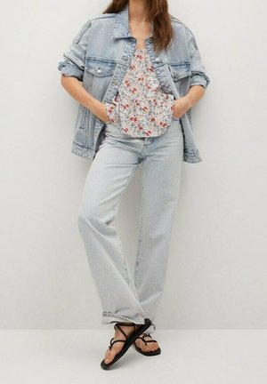 Femme debout les mains dans les poches portant une veste en jean bleu clair, un top fleuri, un jean bleu clair et des sandales noires à brides.