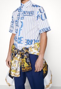 Kortärmad skjorta med knappar i vitt med blå vertikala ränder och grafisk text. Kombinerad med en mönstrad scarf i svart, vitt och guld.