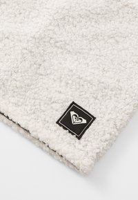 Tissu en polaire texturé blanc avec un patch carré noir portant un logo ; matériau doux et moelleux, forme arrondie et coutures visibles.