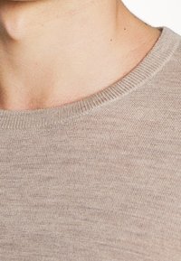 Gros plan sur le cou et l'épaule d'une personne portant un pull en tricot beige à col rond, avec un accent sur la texture du tissu et le détail de l'encolure.